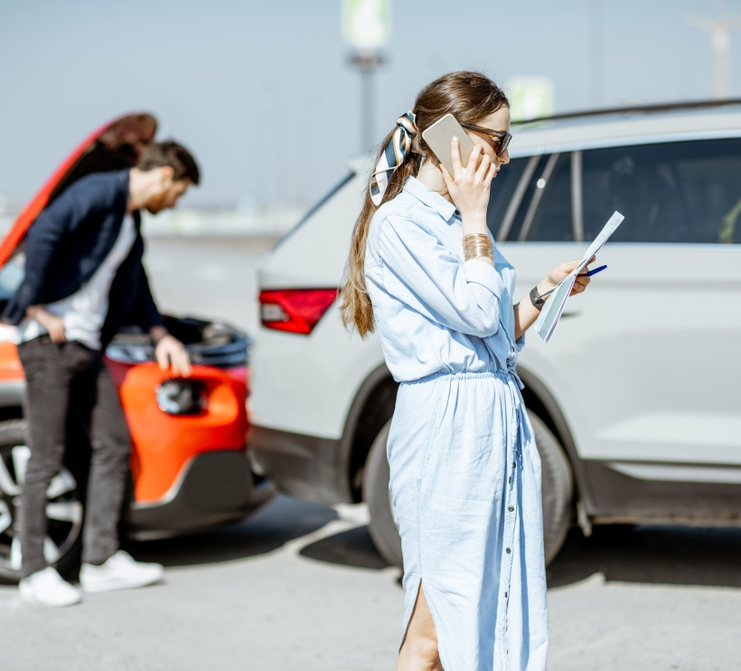 Kobieta rozmawia przez telefon trzymając kartkę papieru, w tle mężczyzna zagląa pod maskępomarańczowego auta, które prawdopodobnie uderzyło w białe auto